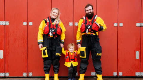Emily Craven, who has blonde hair, and Tom, who has brown hair and a thick broan beard, smile at the camera holding the hand of a toddler with curly blonde hair. All three are wearing yellow and black full body waterproofs and red life-vests. 