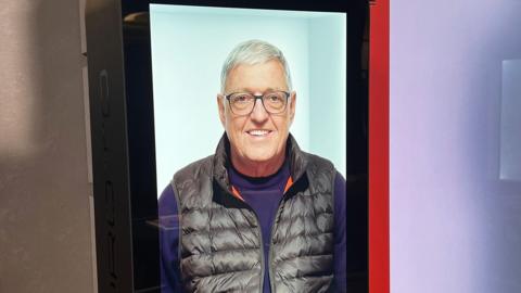 Hologram version of a man called Bill on a large screen, wearing a purple jumper and black gilet and smiling