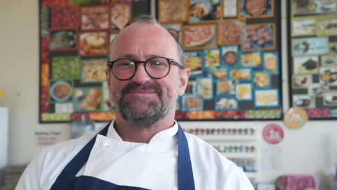Daniel Smith inside a classroom wearing chef whites and a blue apron. He has a pair of black framed circular glasses on and is looking off camera mid-speech.