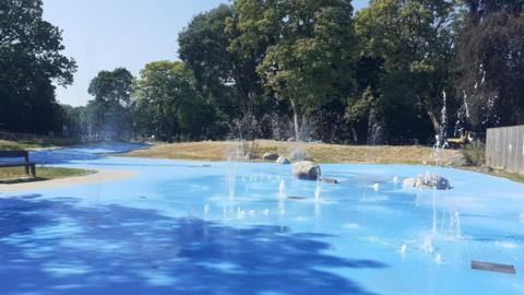 A large, blue water play area with fountains sprouting amid rocks and green trees in the background under a blue sky.