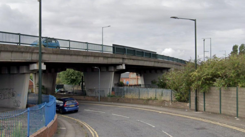 The concrete structure of the Cleethorpe Road bridge taken from a street below. A blue car is travelling over the bridge and another vehicle can be seen travelling underneath.