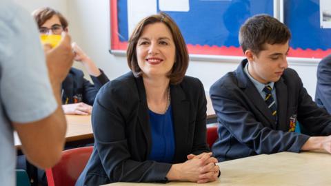 Phillipson wears a black suit jacket over a royal blue dress with a silver necklace. Her hair is worn in a dark brown, chin-length bob. She wears bright red lipstick and is smiling at a teacher, who can partially be seen in the foreground on the left. There are pupils sitting around her, all wearing a navy school uniform with a pastel blue shirt and a navy tie with pale blue and gold diagonal stripes. 