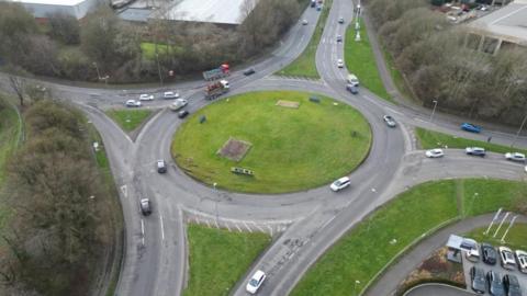 An aerial photo of a roundabout connecting four roads, with a grassy bank in the middle.