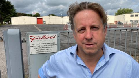 A man in a blue shirt with the collar undone, standing in front of a gate and a large industrial building behind