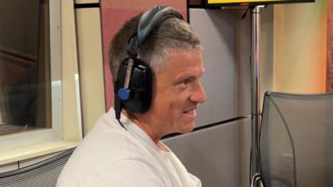 A side profile of Justin Rees, a man with short hair wearing a white T-shirt. He is in a radio studio and wears black headphones, and appears to be talking to someone off camera.