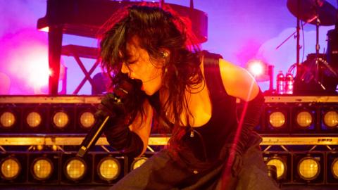 Abigail Morris of The Last Dinner Party performs at O2 Academy Leeds on 24 September. Abigail kneels on stage as she sings into a microphone, her long dark hair worn loose. She wears a black skirt, long black gloves and corset-style top. Behind her there's a piano and drum kit, the stage is lit purple behind her.