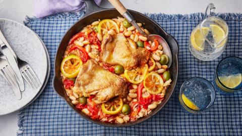 A round pan of Mediterranean chicken with tomatoes, beans, rice, olives and lemon slices is in the middle of the image. It is sitting on a table on top of a blue and white check tablecloth. There is a pile of light blue plates on the left hand side with silver cutlery placed on top, and a jug and two glasses of water with lemon slices in it, to the right.
