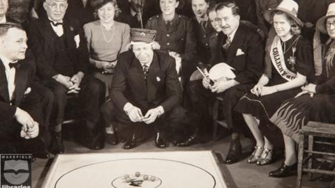 A black and white image of men in suits and women in dresses watching George Formby play marbles