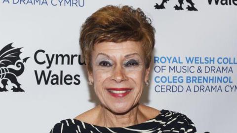 Iris Williams smiling at the camera. She wears a black and white top. She is standing in front of a white background with Cymru Wales, in black writing, and Royal Welsh College of Music & Drama, in blue writing, behind her.