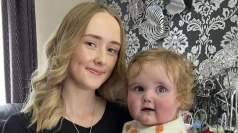 A woman with blonde hair and a black top with a baby with blonde curly hair in front of black and white patterned wallpaper and a grey curtain.