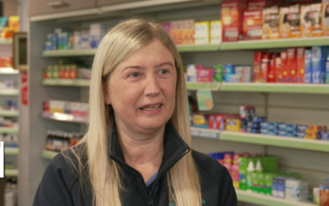 A lady with long blonde hair is talking to the camera and is wearing a black zip-up jacket. Behind her is medication on a shelf.