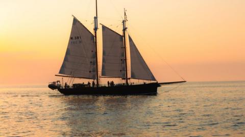 A ship with three white sails in the water. Behind it is an orange skyline.