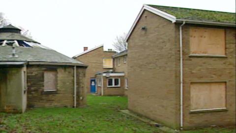 A collection of building boarded up with a blue door in the distance and grass betwen them.