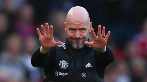 Erik ten Hag, dressed in black Manchester United training gear with white trim, grimaces in the direction of the camera and holds both hands out in front of himself at face level.