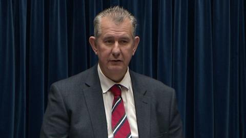 Edwin Poots is wearing a grey suit with a white shirt and a red tie with white and blue slanted stripes. He has short grey hair.