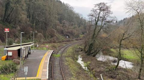 It's a rural railway station with a small station house. You can see the track and to the side of it are some trees where the ground is still holding a lot of water.