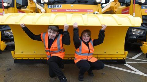 Two school boys wearing hi-vis jackets are crouching under the plough of a large yellow gritter. They have their arms raised and are holding the gritters. A red sign on the gritter announces its name as Ready Salted.