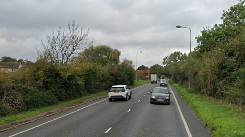 Cars are pictured driving on a road in both direction. The road is lined by a grass verge and hedges. Buildings can be seen in the distance as well as lamp posts.