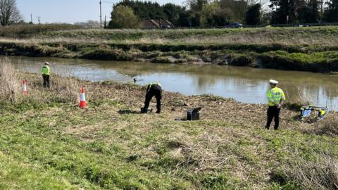 Three police officers are spread out and standing on the side of a river bank. There are two cones on the left and specialist equipment on the grass. A diver is in the water.