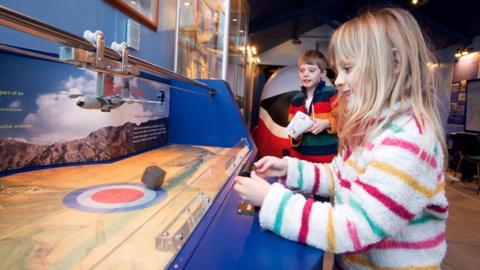 A girl plays with an interactive display about dropping supplies from a RAF aircraft. She has blond shoulder length hair and is wearing a fluffy white top with horizontal multi-coloured strips. A boy with brown hair and striped top watches her in the background