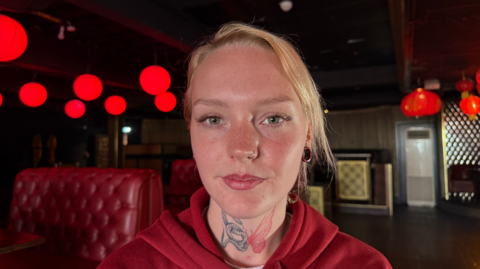 Amber Clarke is sat on a chair looking directly in the camera. She has blonde hair and is wearing a red hoody. She has a tattoo of a snake and butterfly on her neck. Amber is sat on a chair in a nightclub, behind her are glowing red lights which hang from the ceiling as well as red leather seating booths.