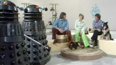 Peter Purves, Lesley Judd, John Noakes, and Shep sit happily in the Blue Peter studio while two Daleks look on.