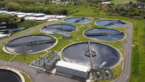 A birdseye view of sewage treatment works. There are six circular containers of water in total, they are laid out as two rows of three pools. The containers are surrounded by grass and there is a separate section in the distance of white containers in an industrial-looking unit. The whole area is surrounded by green fields and trees.