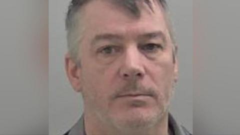 Michael Morris seen in a police mugshot. He has a greying beard and short hair and is looking at the camera.