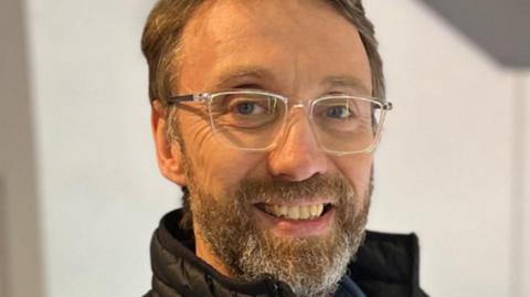 A man with brown hair and a beard with silver-framed glasses and a high black collar