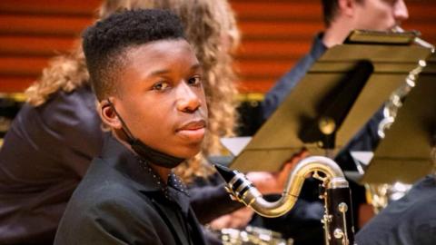 A young black boy sits in a chair and holds a clarinet during a school orchestra performance