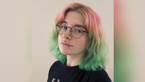 Portrait photo of young woman with pink and green hair, round glasses and a number of lip piercings.