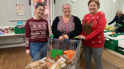 Three volunteers from Cheylesmore Food Hub are standing behind a large shopping trolley full of bread