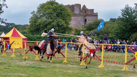 Two knights on horseback clash in a jousting competition if front of a large crowd with trees and a castle behind them