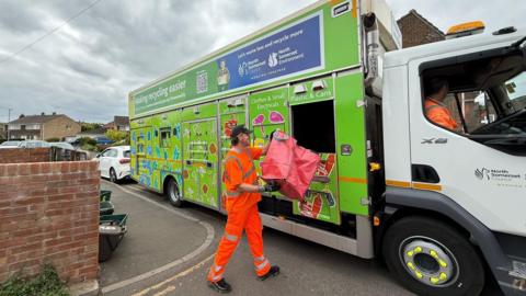 A refuse worker empties one of the new large red plastic recycling bags into a green recycling truck parked on a residential street.