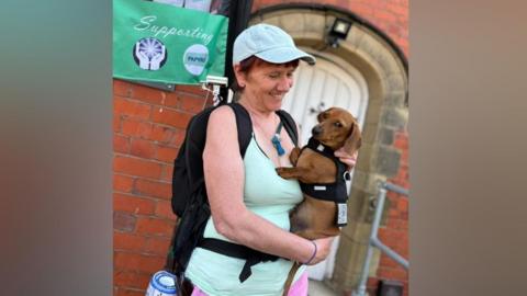 A woman holding a dacshund in her arms. She is wearing sportswear - a pale turquoise vest, a pale blue baseball cap, and a black rucksack from which a collection tin dangles. She is smiling and looking at the dog which is light brown, with a somewhat quizzical expression and wearing a black harness. Behind her is a brick wall, with a white door and part of a green banner can be seen.