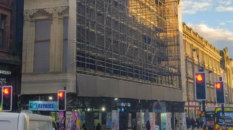 Scaffolding and canvas cover a wide building on a busy street. Traffic waits behind red lights and pedestrians walk on the pavement under the scaffold.