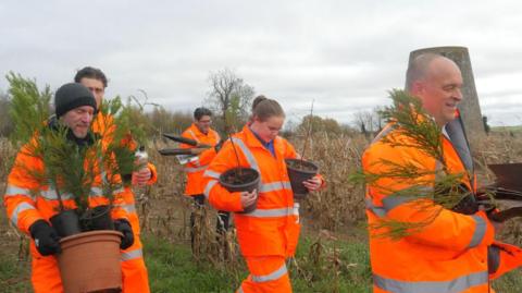 People in orange high vis carrying plant pots with trees in across the countryside