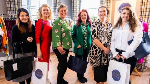 Six women stand in front of clothes rails, holding shopping bags