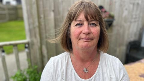 A woman in her 50s is sat on a garden bench with a fence behind her. She has mid length brown hair in a bob hair cut, and is wearing a white top with a silver necklace. She has a passive expression. 