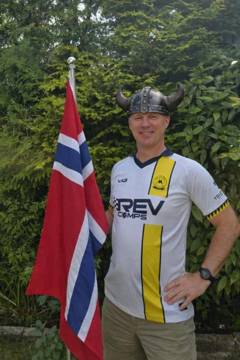 Man in a white and yellow Torquay United football top wears a Viking helmet and holds a red and blue and white Norweigan flag in front of a green hedge