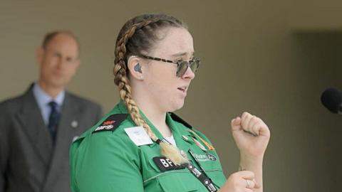 Hafwen Clarke wearing a green St John Ambulance shirt and her blonde hair is in pigtails, her hands are up as she is signing