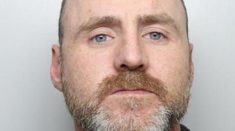 A man staring straight ahead. He has a ginger and grey-coloured beard with very short hair.