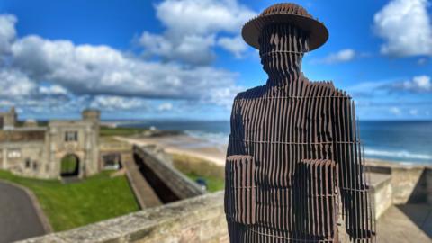 Sculpture of soldier constructed from vertical steel plates standing on a castle wall with the sea in the background.