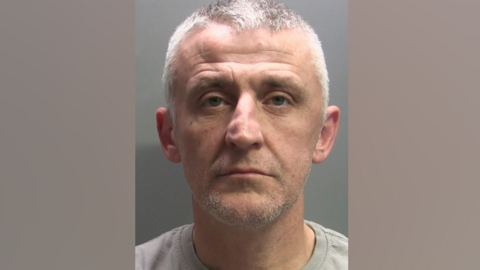 A mugshot of Stephen Young who is looking into the camera with a slight frown. He has grey, short hair and a small face tattoo underneath his right eye. He is wearing a grey t-shirt.