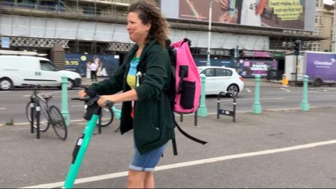 A woman, Shona Mackintosh, riding a turquoise e-scooter on a seafront cycle lane
