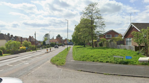 A straight road taken from a small junction. A grass bank runs down the length of the road on the right of the road with houses either side of it. 