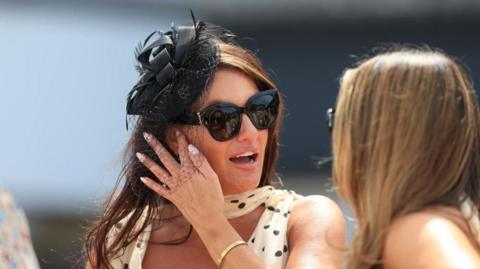A woman with dark hair wears a white and black formal polka-dot dress, with a black fascinator and large black sunglasses - she is talking to a woman with blonde hair, and both are outside 