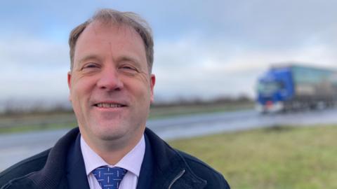 Ashley Baxter stands at the side of a road as a lorry drives past. He has thinning hair and is wearing a blue coloured jacket, lilac shirt and a blue tie.