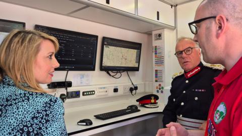 Interior of a control room inside a mobile or modular unit, with two wall-mounted computer screens displaying search-and-rescue software, including a digital map with marked locations and a status dashboard. Two men and a woman are in vision.