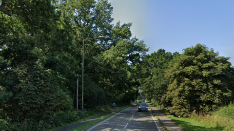 A google street view image of a country road, showing trees and woodlands on either side of a road with two lanes. A car is driving towards the viewer